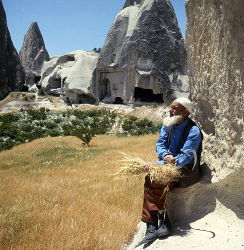 Farmer with sheaf of oats in front of cone dwellings, Cappadocia, Turkey